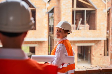 Photo for Smiling construction manager with rolled-up architectural drawing under her arm looking at contractor while standing in front of unfinished houses - Royalty Free Image