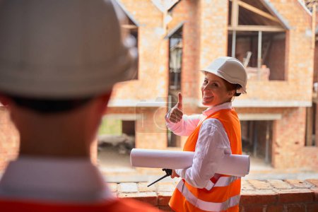 Photo for Joyful building supervisor with rolled-up blueprints under arm giving thumbs-up to foreman while inspecting unfinished private residential complex - Royalty Free Image