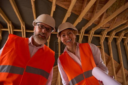 Téléchargez les photos : Entrepreneur et inspecteur de bâtiment joyeux avec des plans roulés à la main debout sous des fermes de toit en bois dans le grenier - en image libre de droit