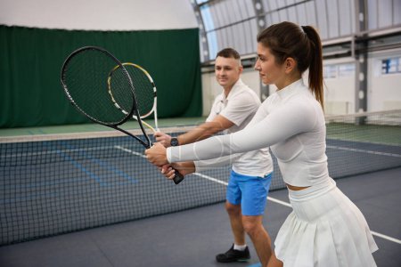 Téléchargez les photos : Homme sportif instructeur enseigne à la femme qu'ils jouent au tennis travail sur frapper sur le terrain intérieur - en image libre de droit