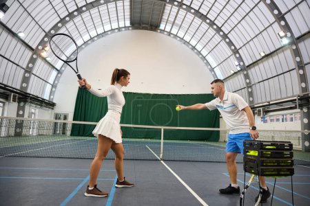 Téléchargez les photos : Instructeur de longueur complète de l'homme aide la femme à apprendre le tennis qu'ils pratiquent frapper la balle sur le terrain intérieur - en image libre de droit