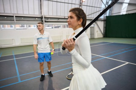 Téléchargez les photos : Homme instructeur aide femme apprendre le tennis ils pratiquent frapper balle sur le terrain intérieur - en image libre de droit