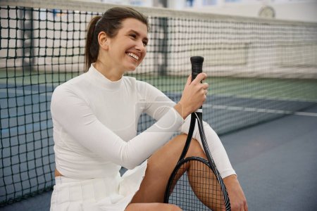 Photo for Smiling cheerful woman with racket in hands sitting on tennis court - Royalty Free Image