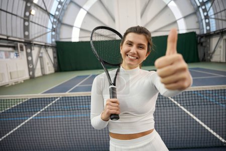 Téléchargez les photos : Femme en forme satisfaite en tenue blanche avec raquette debout montrant pouce vers le haut sur le court de tennis intérieur - en image libre de droit