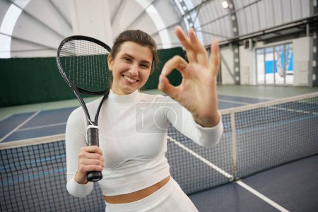 Téléchargez les photos : Femme sportive en tenue blanche avec raquette debout montrant ok signe sur le court de tennis intérieur - en image libre de droit