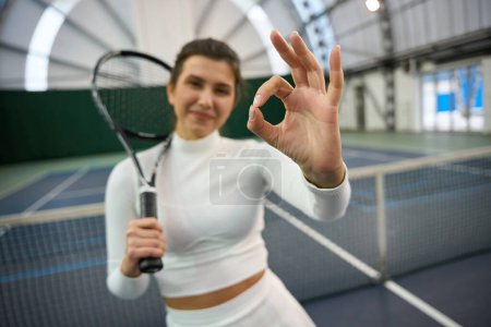 Foto de Atlética mujer sonriente en traje blanco con raqueta de pie mostrando buen gesto en la cancha de tenis cubierta - Imagen libre de derechos