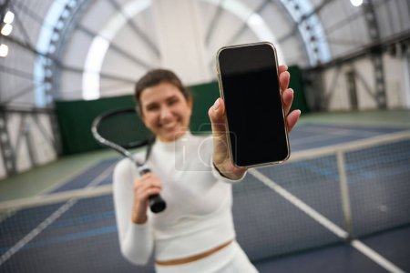 Téléchargez les photos : Joueuse en forme souriante montrant un écran de téléphone vierge sur un court de tennis intérieur - en image libre de droit