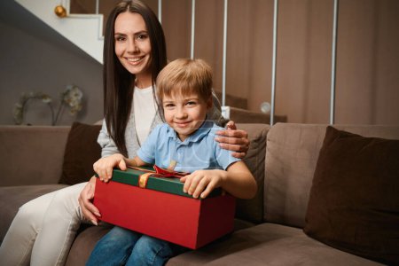 Photo for Smiling woman sitting with her son giving New Year gift celebrating Christmas at home - Royalty Free Image