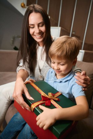 Foto de Encantada madre presentando regalo de Navidad a su hijo en medio de las festividades de vacaciones de invierno en el hotel - Imagen libre de derechos
