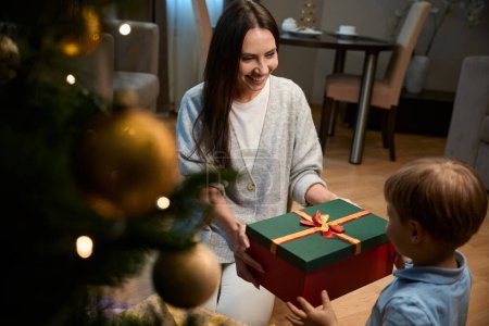 Photo for Cheerful woman and her son sharing festive spirit of Christmas exchanging presents beside the holiday tree in hotel - Royalty Free Image