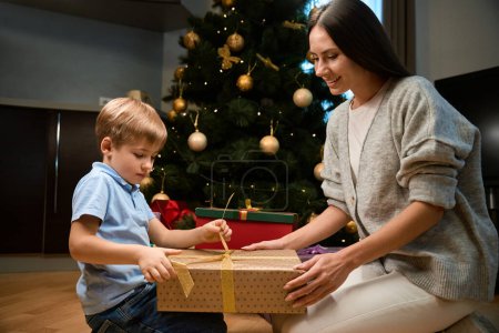 Foto de Encantada madre y su hijo intercambian regalos de Navidad rodeados por el brillo del árbol de Navidad en el hotel - Imagen libre de derechos