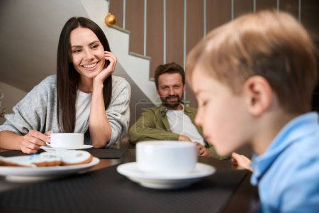 Téléchargez les photos : Femme souriante regardant son fils tout en prenant le petit déjeuner de vacances dans la chambre d'hôtel - en image libre de droit