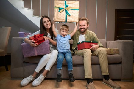 Téléchargez les photos : Parents avec petit garçon tenant des boîtes-cadeaux célébrant les vacances du Nouvel An à l'hôtel tout en étant assis ensemble sur le canapé - en image libre de droit