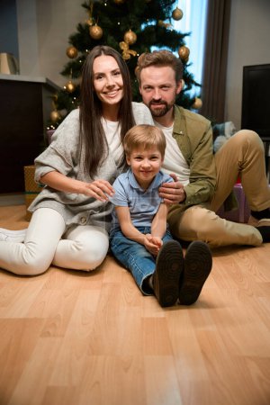 Foto de Familia sonriente sentada en el piso cerca del árbol de Navidad decorado en el hotel celebrando el Año Nuevo juntos - Imagen libre de derechos