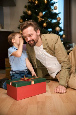 Foto de Niño compartiendo secreto con su padre mientras está sentado cerca del árbol de Navidad y la caja de regalo celebrando las vacaciones de invierno juntos - Imagen libre de derechos