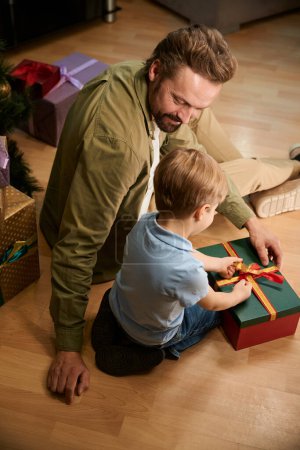 Photo for Man and little boy sitting on floor opening Christmas present celebrating New Year at home - Royalty Free Image