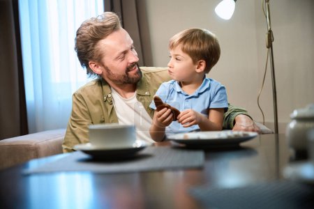 Téléchargez les photos : Petit garçon assis à la table avec père ayant du pain d'épice pour le petit déjeuner de Noël à l'hôtel - en image libre de droit