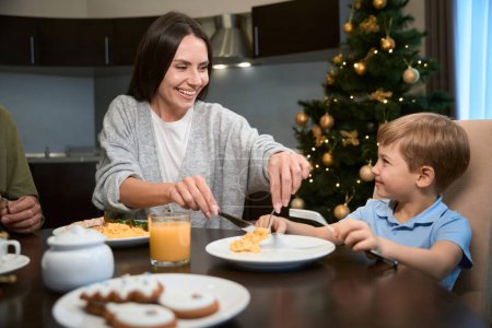 Photo for Cheerful woman having dinner with little boy relaxing during festive lazy breakfast during New Year celebration - Royalty Free Image