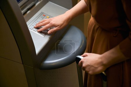 Téléchargez les photos : Image recadrée de la femme avec valise appuyant sur le bouton sur ATM dans le hall de l'hôtel. Concept de repos, vacances et voyages - en image libre de droit