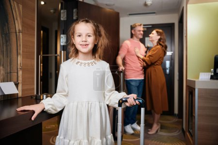 Photo for Smiling little girl with suitcase looking at camera with blurred mother and father on background in hotel room. Rest, vacation and travelling. Idea of family relationship and spending time together - Royalty Free Image