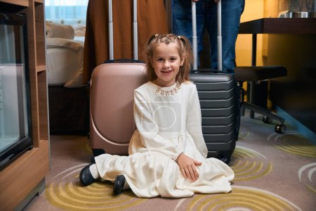 Téléchargez les photos : Petite fille souriante assise sur le sol et regardant la caméra près de la mère et du père recadrés avec des valises sur fond dans la chambre d'hôtel. Repos, vacances et voyages. Relations familiales - en image libre de droit