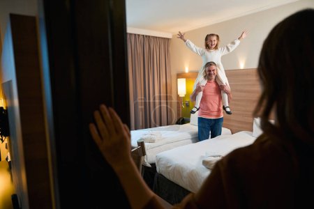Photo for Cropped mother looking at joyful daughter with hands in air on fathers shoulders in hotel room. Concept of rest, vacation and travelling. Idea of family relationship and spending time together - Royalty Free Image
