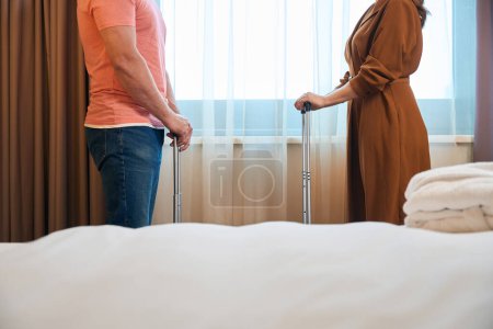 Photo for Cropped side view of couple with suitcases between bed and window in hotel room. Concept of rest, vacation and travelling - Royalty Free Image