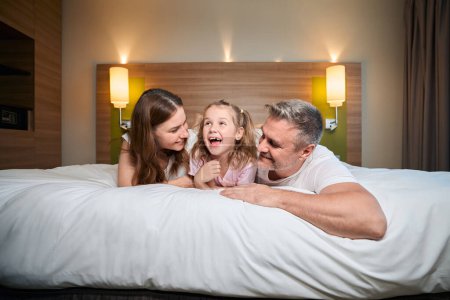 Photo for Happy european family of mother, father and joyful daughter embracing while lying on bed in hotel room. Concept of rest, vacation and travelling. Idea of family relationship and spending time together - Royalty Free Image