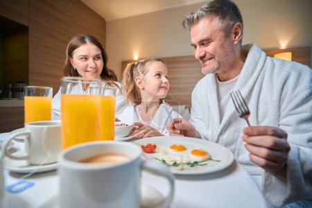 Téléchargez les photos : Mère et fille regardant le père pendant le petit déjeuner sur le lit à table dans la chambre d'hôtel. Concept de repos, vacances et voyages. Idée de relation familiale et de passer du temps ensemble - en image libre de droit