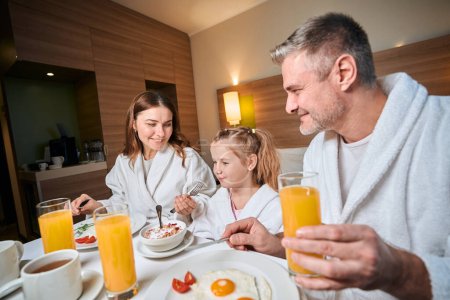 Téléchargez les photos : Père regardant la mère nourrir sa fille flocons pendant le petit déjeuner dans la chambre d'hôtel. Concept de repos, vacances et voyages. Idée de relation familiale et de passer du temps ensemble - en image libre de droit