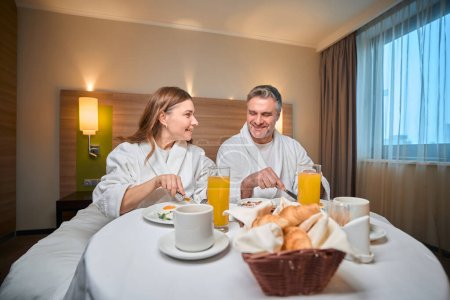 Téléchargez les photos : Couple caucasien adulte souriant portant peignoir petit déjeuner sur le lit à la table dans la chambre d'hôtel. Concept de repos, vacances et voyages - en image libre de droit