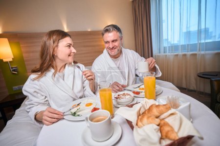 Foto de Pareja caucásica adulta sonriente que usa albornoz desayunando y mirándose en la cama a la mesa en la habitación del hotel. Concepto de descanso, vacaciones y viajes - Imagen libre de derechos