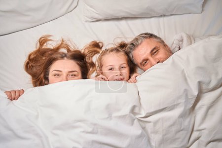 Photo for Top view of caucasian mother, father and daughter covering face with blanket and looking at camera on bed in hotel room. Rest, vacation and travelling. Family relationship and enjoying time together - Royalty Free Image