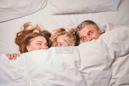 Photo for Top view of caucasian family of mother, father and daughter covering face with blanket and looking at each other on bed in hotel room. Rest, vacation and travelling. Family relationship - Royalty Free Image