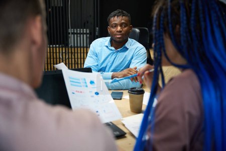 Foto de Líder de la compañía afroamericana escuchando el informe de sus colegas caucásicos, equipo discutiendo estrategia de negocios y recopilación de datos de mercado en la reunión en la oficina - Imagen libre de derechos