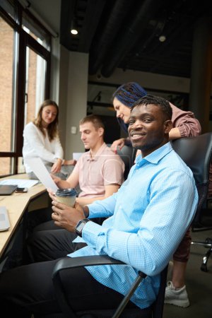 Téléchargez les photos : Un homme souriant gestionnaire d'une entreprise en développement regardant la caméra assise sur le lieu de travail avec d'autres collègues, une équipe multiethnique discutant du processus de travail, partageant des idées - en image libre de droit