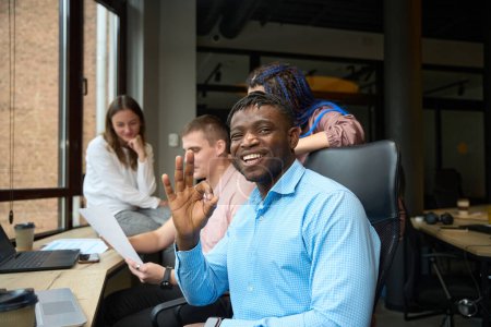 Téléchargez les photos : Homme afro-américain joyeux montrant un geste correct de la main et regardant la caméra avec un sourire dents, travaillant en compagnie multiethnique avec un personnel amical - en image libre de droit
