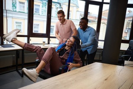 Foto de Mujer sonriente con el pelo trenzado azul disfrutando de dos hombres multiétnicos rodando ella en sillón alrededor de la oficina, equipo de gerentes divirtiéndose y jugando durante el descanso, tradiciones de la oficina - Imagen libre de derechos