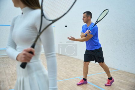 Téléchargez les photos : Enthousiaste homme entraîneur enseignant à la femme des compétences précises de raquette dans le squash sur cour intérieure - en image libre de droit