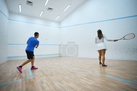 Téléchargez les photos : Femme motivée athlète raffine les techniques de frappe de squash sous l'homme instructeurs conseils sur cour intérieure - en image libre de droit