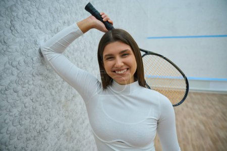 Foto de Atlética mujer alegre sosteniendo raqueta de squash en ella en la cancha de interior - Imagen libre de derechos