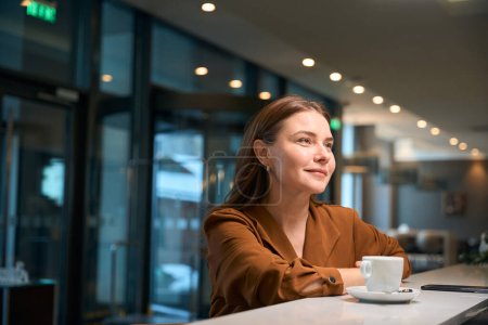 Photo for Adult smiling caucasian woman looking away while drinking tea or coffee at reception desk in hotel lobby. Concept of rest, vacation and travelling - Royalty Free Image