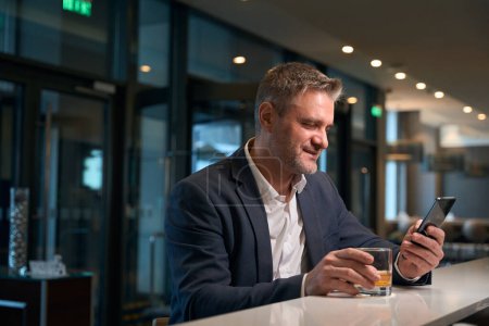 Photo for Adult focused caucasian man using smartphone while drinking cognac from glass cup at reception desk in hotel lobby. Concept of rest, vacation and travelling - Royalty Free Image