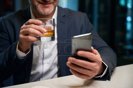 Téléchargez les photos : Visage obscur d'un homme d'affaires adulte utilisant un smartphone tout en buvant du cognac dans une tasse en verre à la réception dans le hall de l'hôtel. Concept de repos, vacances et voyages - en image libre de droit