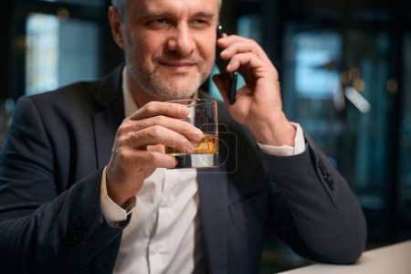 Photo for Partial adult focused caucasian entrepreneur talking on smartphone while drinking whiskey from glass cup at reception desk in hotel lobby. Concept of rest, vacation and travelling - Royalty Free Image