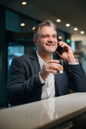 Photo for Adult smiling caucasian man talking on mobile phone while drinking whiskey from glass cup at reception desk in hotel lobby. Concept of rest, vacation and travelling - Royalty Free Image