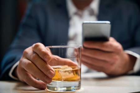 Photo for Focus on foreground of glass cup with cognac or whiskey in hands of blurred businessman using smartphone at reception desk in hotel lobby. Concept of rest, vacation and travelling - Royalty Free Image