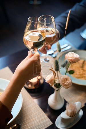 Photo for Partial image of couple toasting wine from glasses at table in hotel restaurant. Concept of romantic date and event - Royalty Free Image