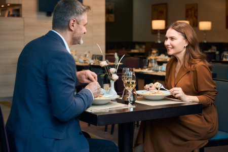 Photo for Adult smiling caucasian couple eating pasta and looking at each other in hotel restaurant. Concept of romantic date and event - Royalty Free Image