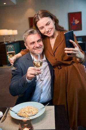 Téléchargez les photos : Couple caucasien souriant adulte avec prise de vin selfie sur smartphone dans le restaurant de l'hôtel. Concept de date et d'événement romantique - en image libre de droit